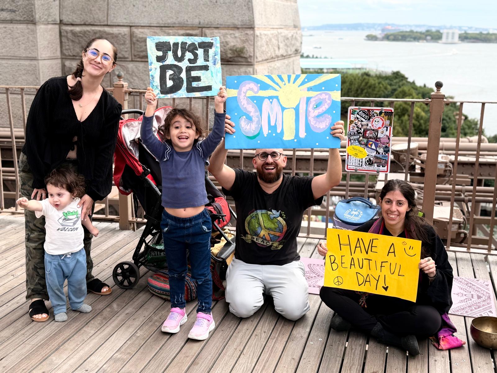 Adults and children together on Brooklyn Bridge holding handmade Just Be, Smile, and Have a Beautiful Day signs